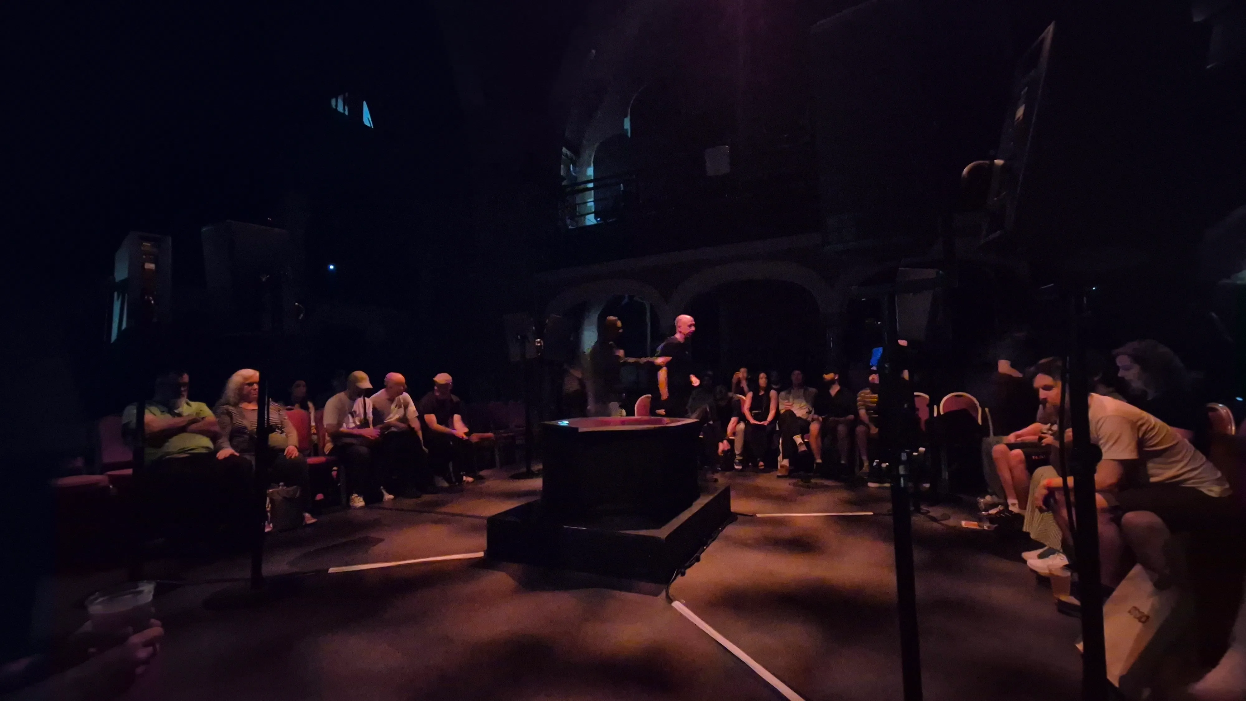 A low-light, eye-level view of an audience gathered in an arched venue. People are seated in a large circle of chairs around a dark, unlit hexagonal structure with a red cover in the center of the room. A few people are standing near the center, and the overall atmosphere is dark and moody.