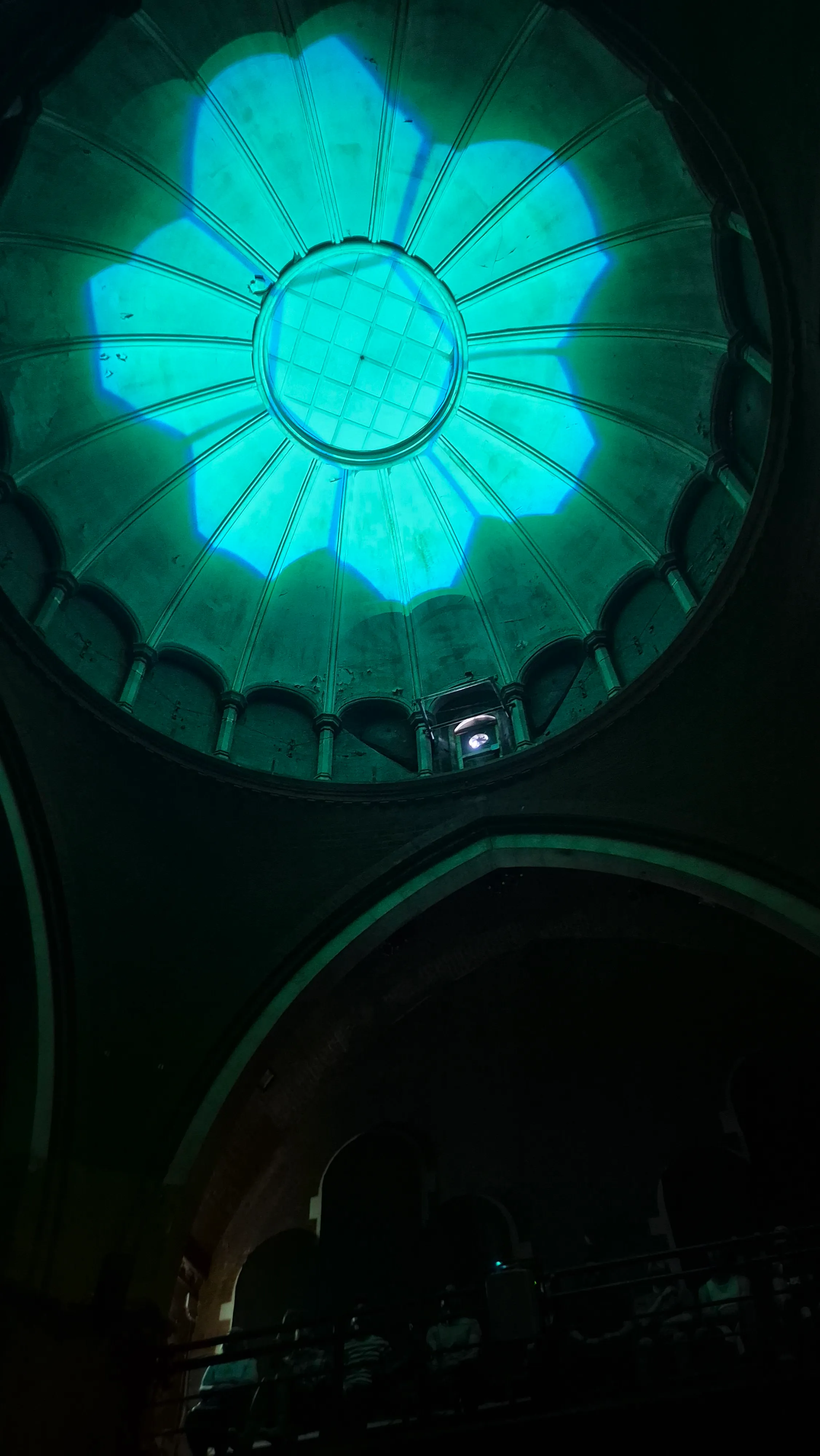 A low-angle shot looking directly up at a classic domed ceiling with architectural ribbing. The dome is illuminated by a large, bright teal light projection shaped like a group of seven hexagons. The dark silhouette of a balcony with seated attendees is visible at the bottom of the frame.
