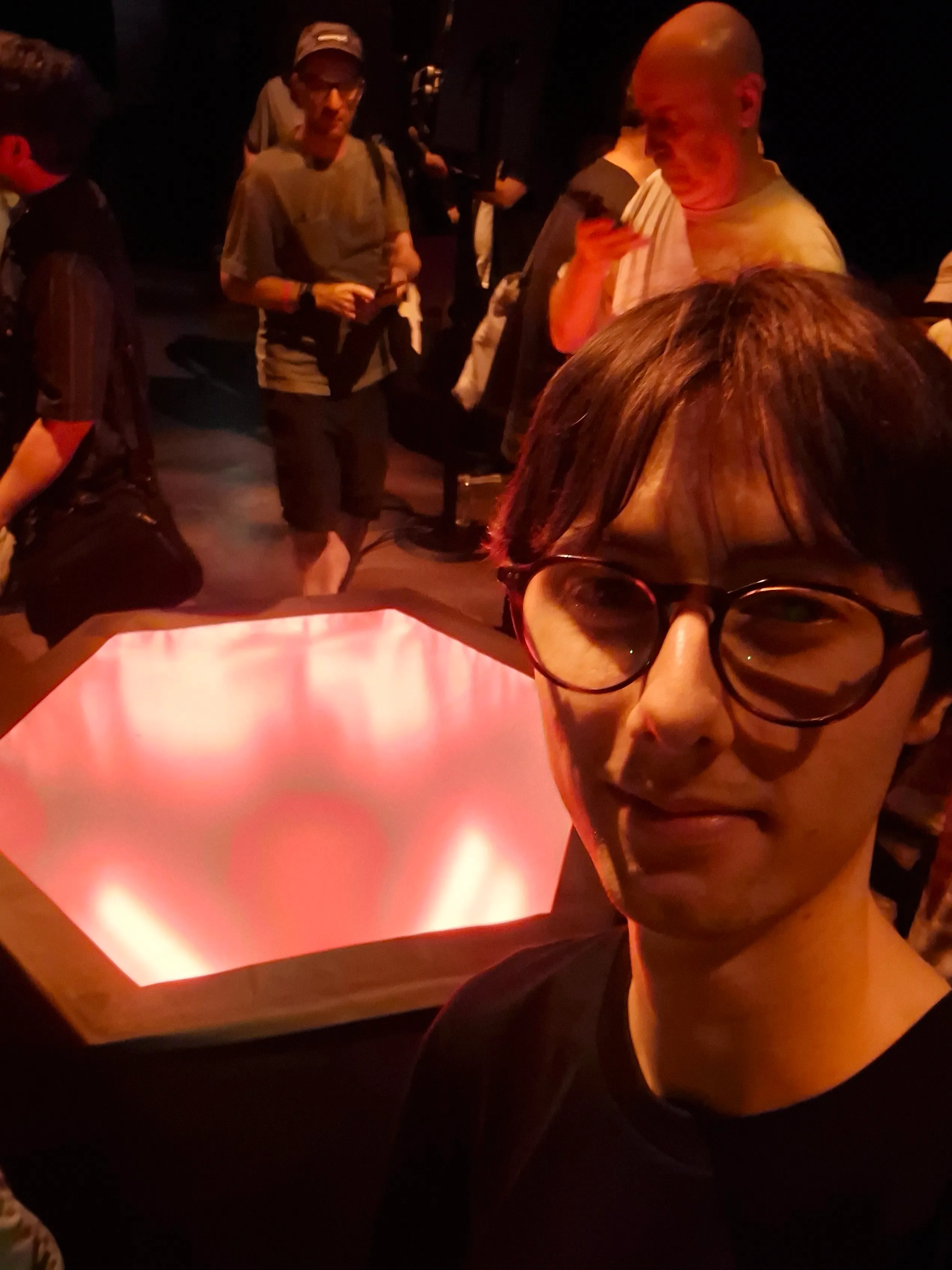 A selfie of a person with dark hair and round glasses. They are standing in a dark room right in front of a hexagonal structure emitting a bright, glowing red light. Other attendees are visible standing in the blurred background.