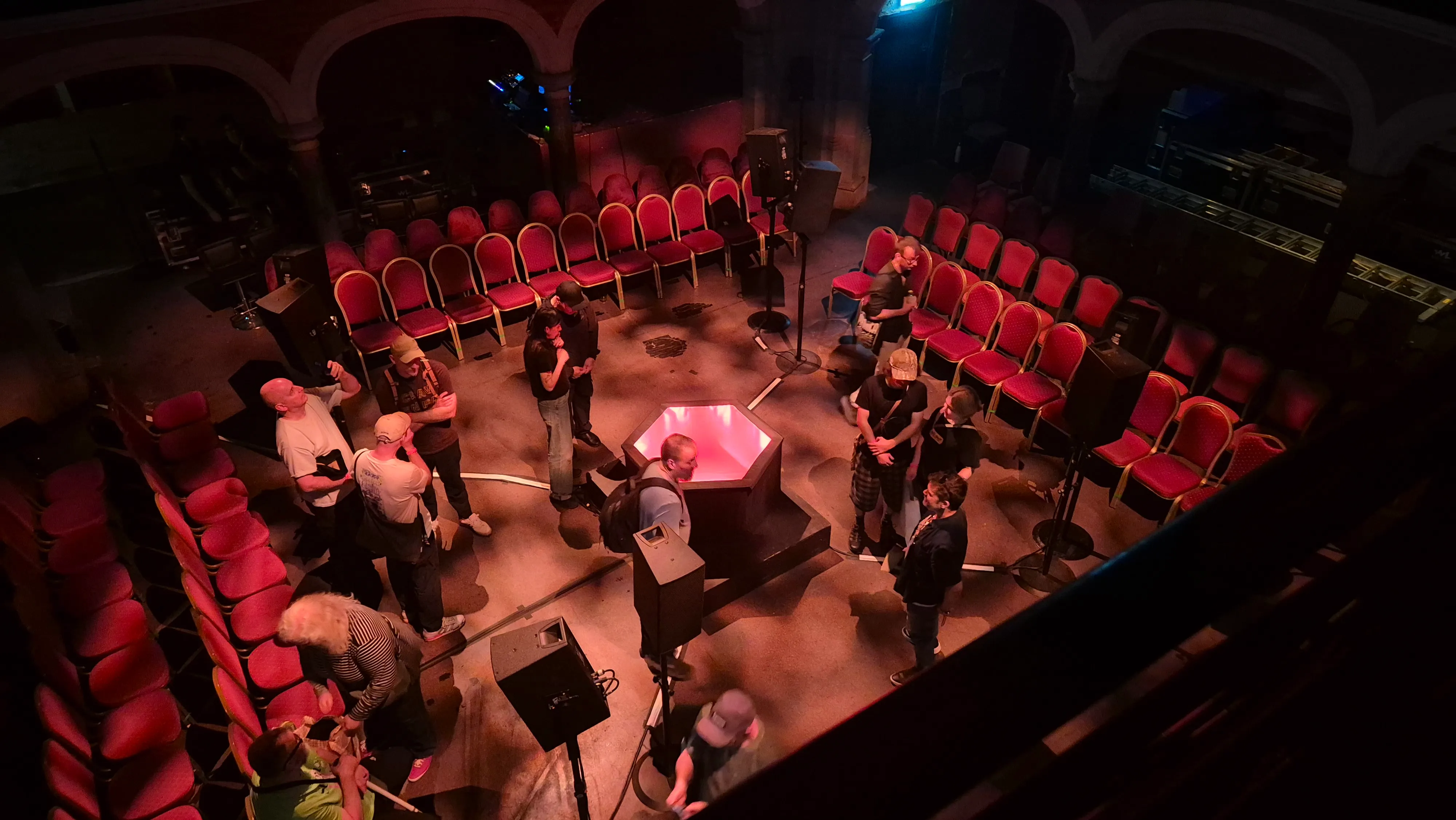 An overhead view of a dimly lit event space characterized by arched alcoves. Rows of red chairs are arranged in a large circle surrounding a central, hexagonal structure that is glowing with red light. Several people are standing and conversing in the center of the circle around the structure.