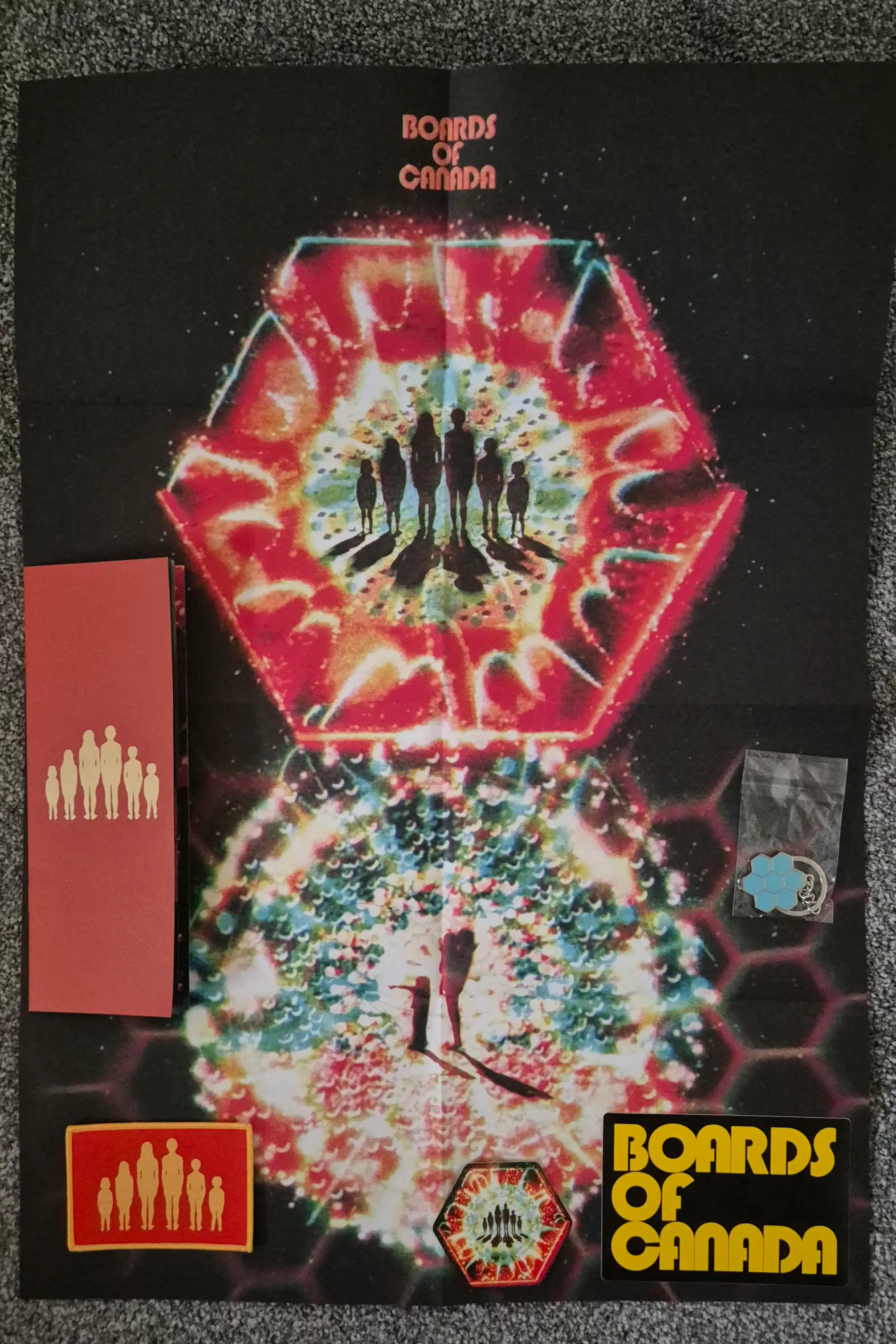 A collection of Boards of Canada merchandise laid out on a gray carpet. The items include a folded poster featuring kaleidoscopic artwork and the band's logo, a tall red pamphlet with an embossed golden silhouette of a family, a red rectangular patch with the same family silhouette, a small hexagonal sticker, a yellow rectangular sticker with the band's logo, and a small light blue hexagonal pin.